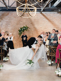 Bride and groom dip kiss at the end of wedding ceremony with guests applauding.