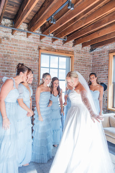 Bride revealing her wedding dress to bridesmaids in matching dusty blue gowns.