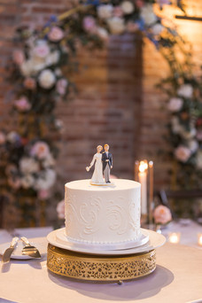 Elegant white wedding cake with bride and groom topper on gold stand in romantic candlelit reception setting.