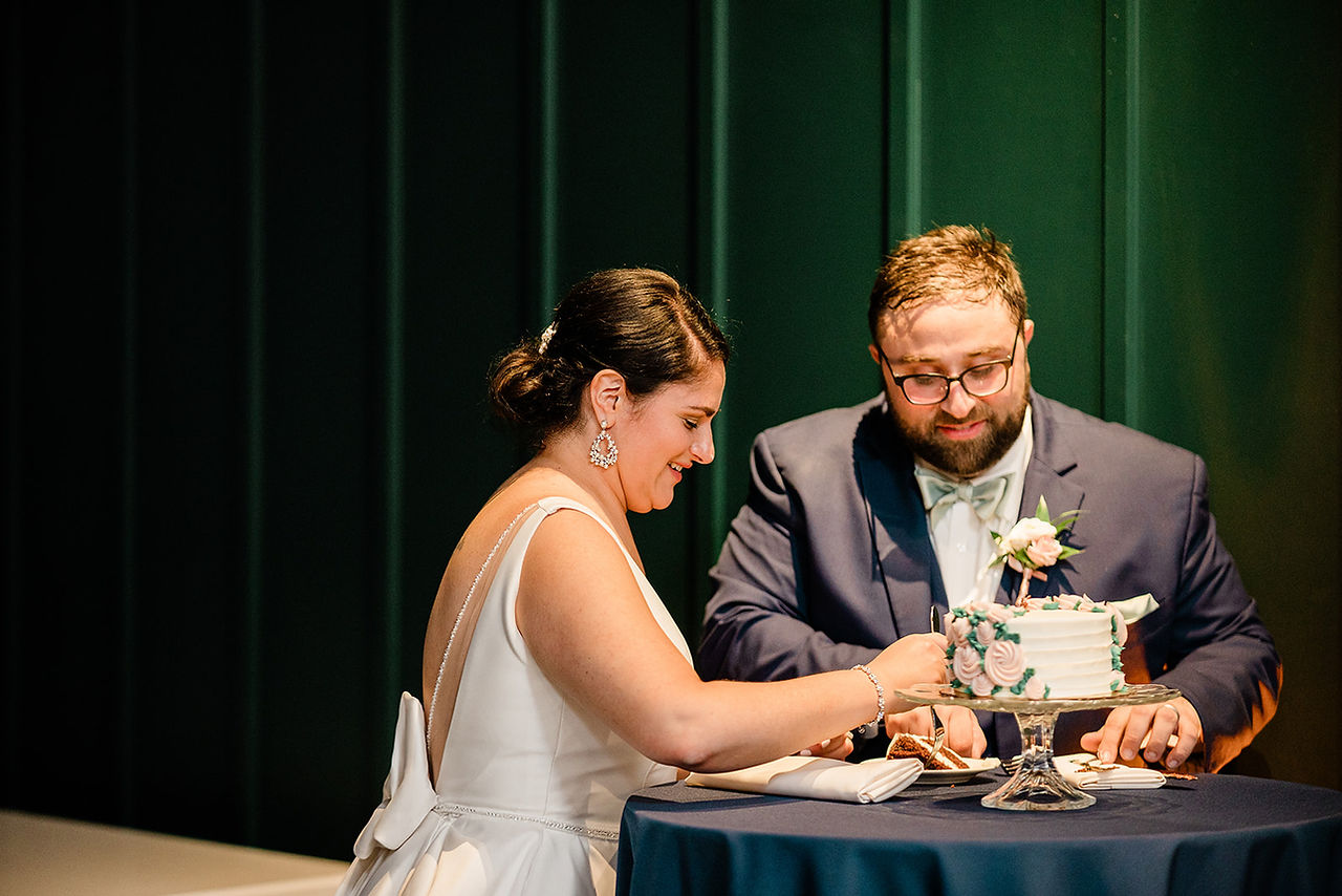 Bride and groom cut wedding cake with green backdrop.