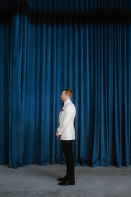 Groom in white tuxedo jacket waiting in front of deep blue velvet curtain before wedding first look.
