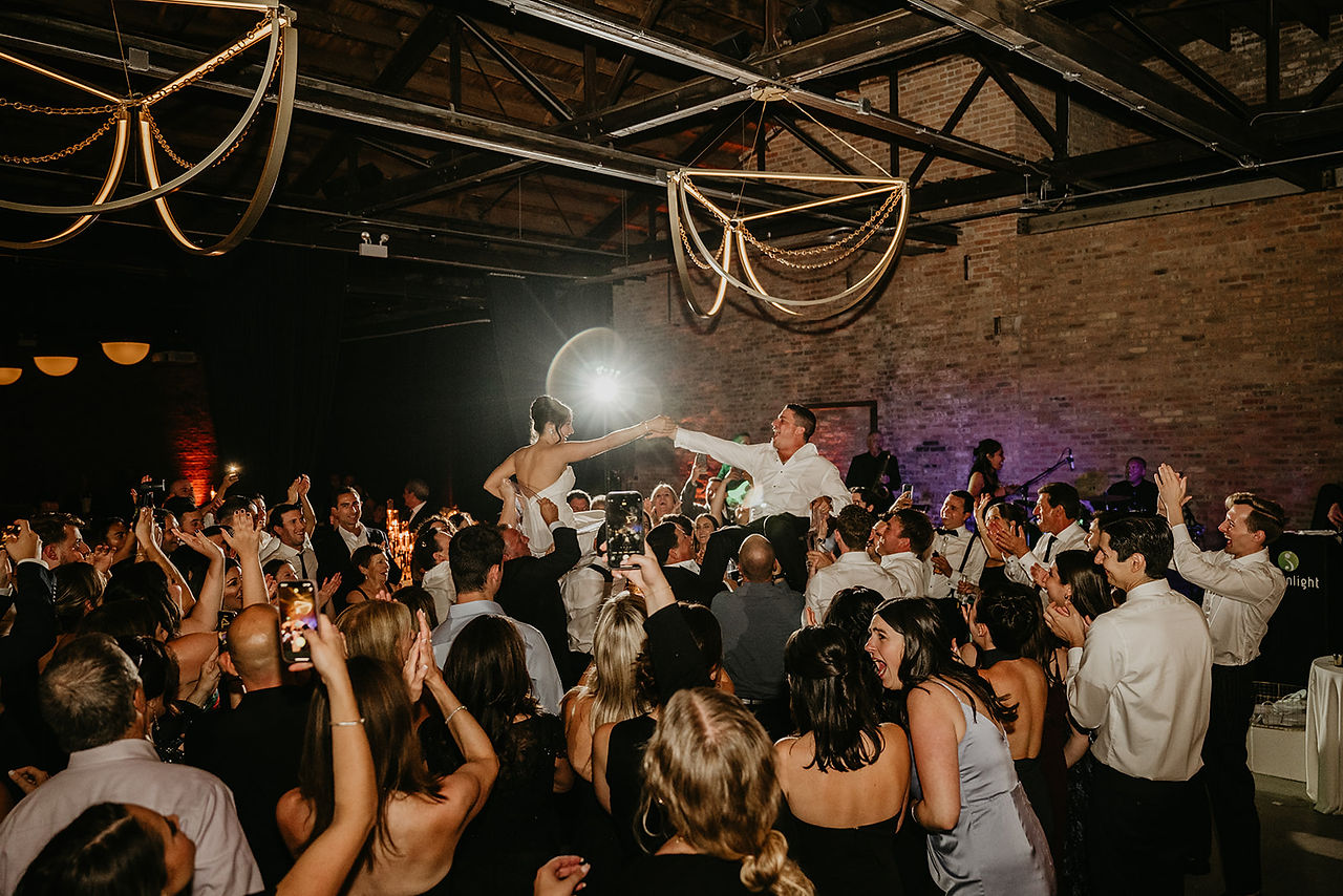 Bride and groom lifted on chairs beneath chandeliers and dancing guests.