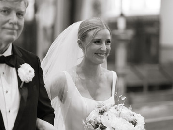 Smiling bride walking down aisle with veil and bouquet in black and white.