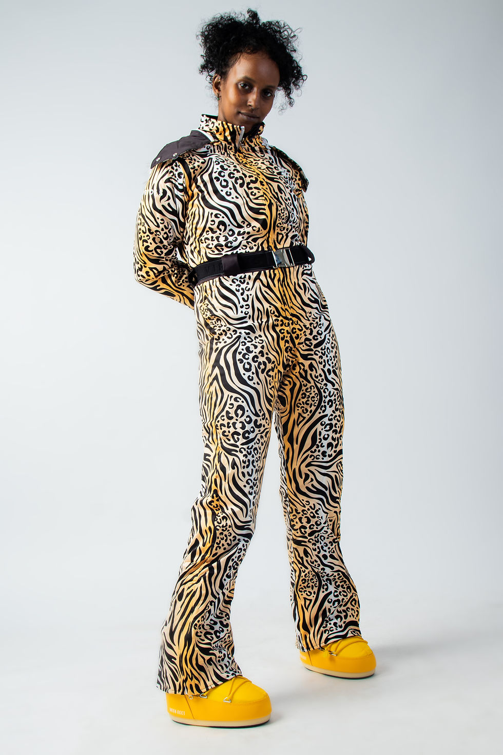Thumbnail: Model wearing the patterned side of the 'Black Piste' reversible ski suit, featuring a black zebra and leopard print with a yellow and white gradient background, standing with legs apart, facing slightly inward, and arms behind her back.