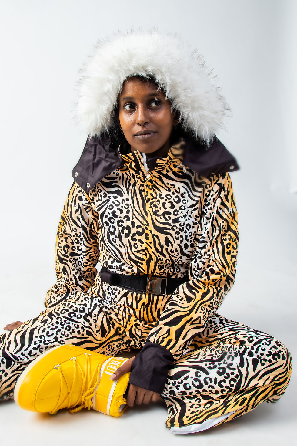 Thumbnail: Model wearing the patterned side of the 'Black Piste' reversible ski suit, featuring a black zebra and leopard print with a yellow and white gradient background, sitting with one leg bent.
