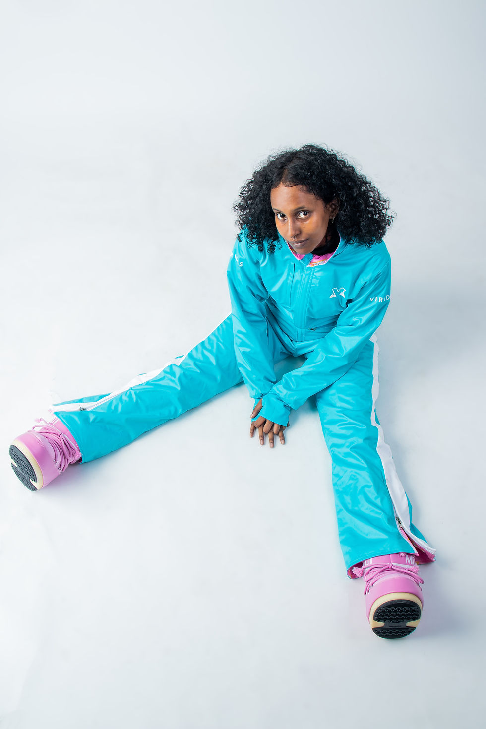 Thumbnail: Model wearing the plain bright blue side of the 'Blue Piste' reversible ski suit, sitting with legs stretched out.