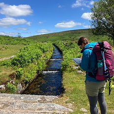 There is a woman with her back to us carrying a rucksack and looking at a map, in front of her is a manmade waterway, a leat, running a head. On either side of the leat is green grass and there is a tree above her head, the sky is blue in the distance