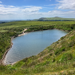Crazywell pool sits in the middle of dartmoor with high sides and one lower side with a small beach. In the distance are other tors and blue skies. The pool is surrounded by the grassy moorland.