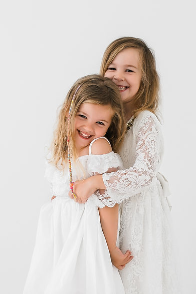 Two young sisters in white dresses hugging and laughing together.