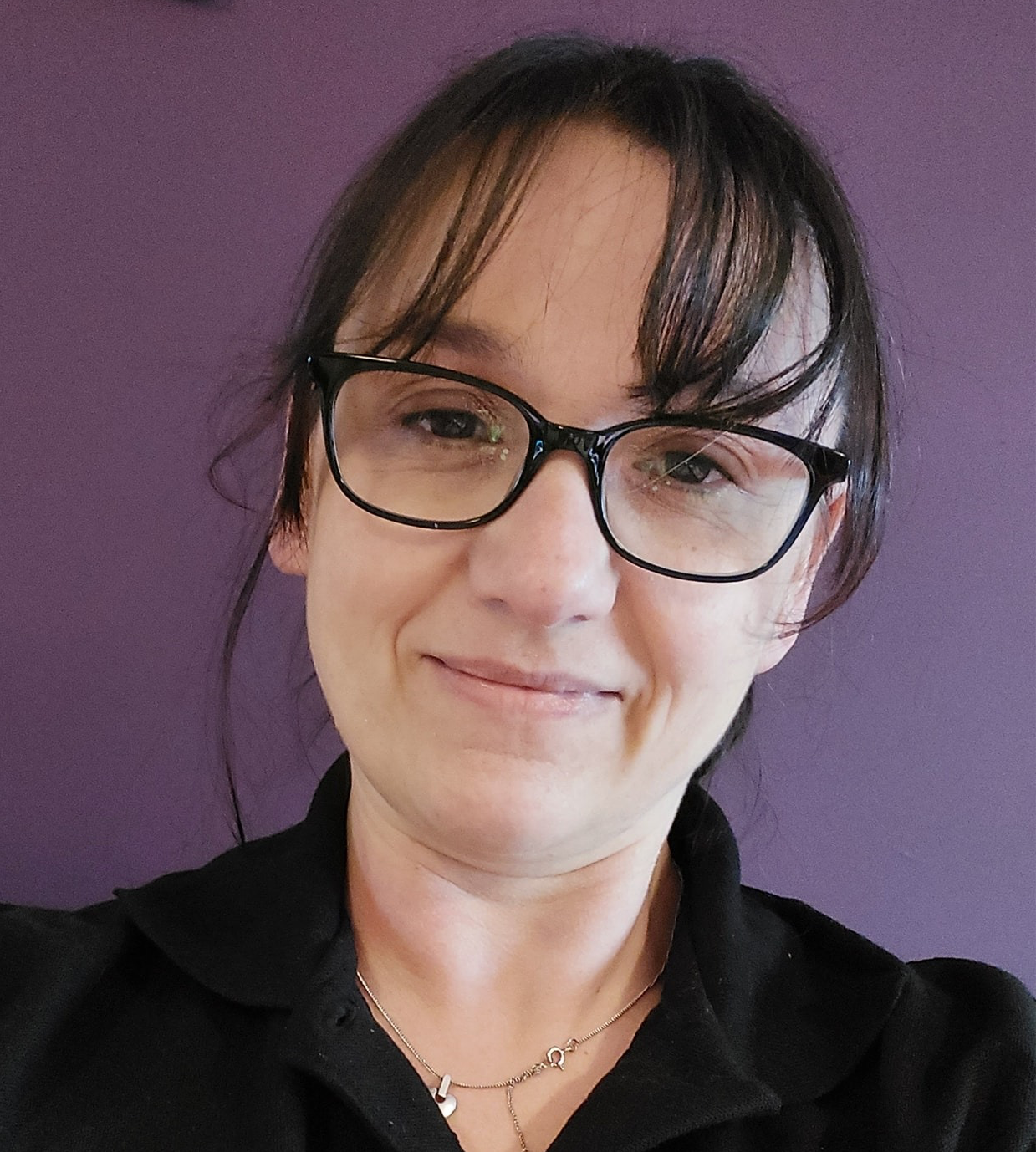 Karen the lactation consultant. She has dark hair, tied back with a fringe. She is wearing a black polo shirt and has glasses. she is in front of a purple background
