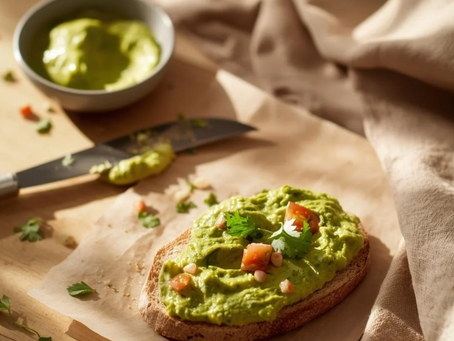 Een snede volkorenbrood belegd met romige guacamole, afgewerkt met stukjes tomaat en verse koriander. Op de achtergrond een kommetje met extra spread, een mes en een linnen doek in zacht ochtendlicht — een natuurlijke en huiselijke sfeer.