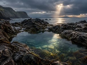 Sunlight breaking through storm clouds over a rocky Irish coastal pool, representing the depth and insight of Mercury in Scorpio.