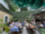A festive outdoor courtyard with guests seated at long tables covered in blue-striped cloths, enjoying an Argentine asado experience while a grill master prepares meat over a wood-fired parrilla. Green balloons hang overhead, and the space is lush with plants and ivy-covered walls, creating a vibrant and welcoming atmosphere.