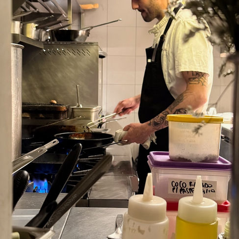 Chef cooking in Alcanfor’s open kitchen in Villa Crespo, Buenos Aires, with herbs and utensils in the foreground.