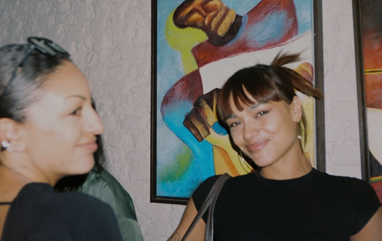 Two women smiling in front of framed paintings at an art gallery.