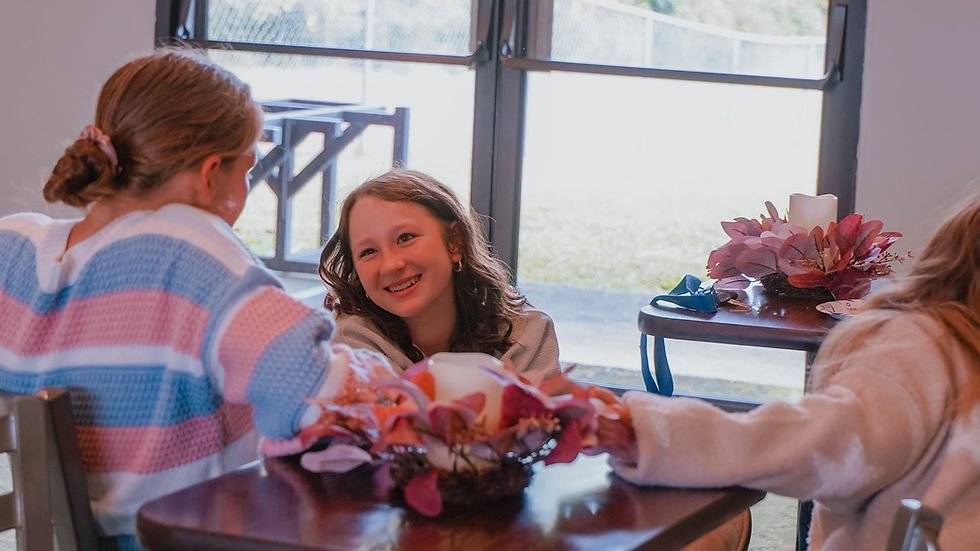 Three young girls laughing closely around a floral arrangement on table.
