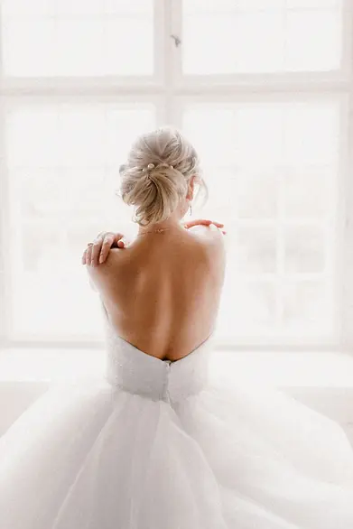 Back view of bride in strapless tulle ballgown adjusting her shoulders in front of a bright window.