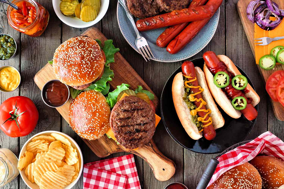 Burgers, hot dogs with mustard and jalapeños, chips, and condiments are arranged on a wooden table, evoking a festive picnic mood.