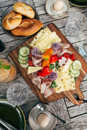 Ein gedeckter Tisch mit einem Holzbrett in der Mitte auf dem Käse, Wurst und Gemüse serviert ist. 