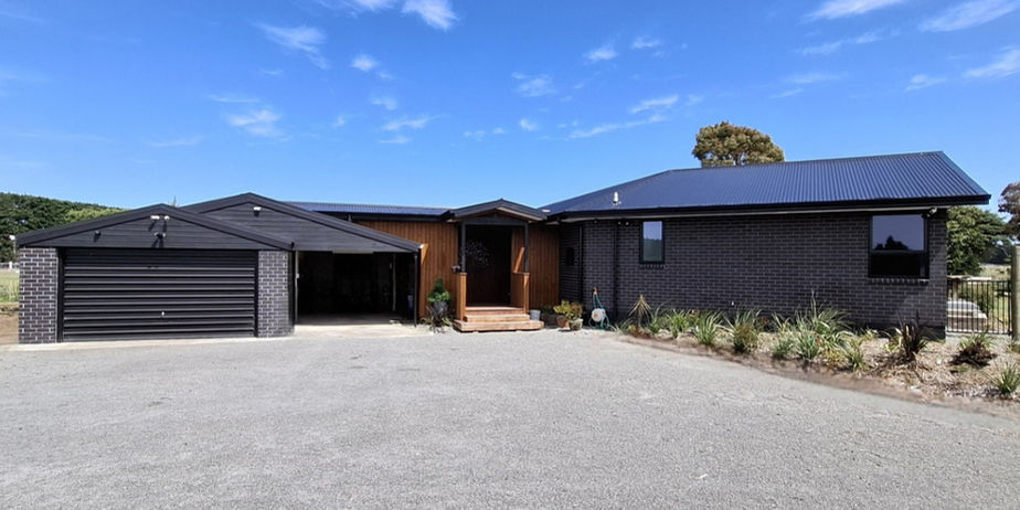 Front exterior of a contemporary single-story home featuring dark charcoal brickwork and a black corrugated metal roof. The design includes a double garage with a black roller door and a warm, vertical timber-clad recessed entryway.