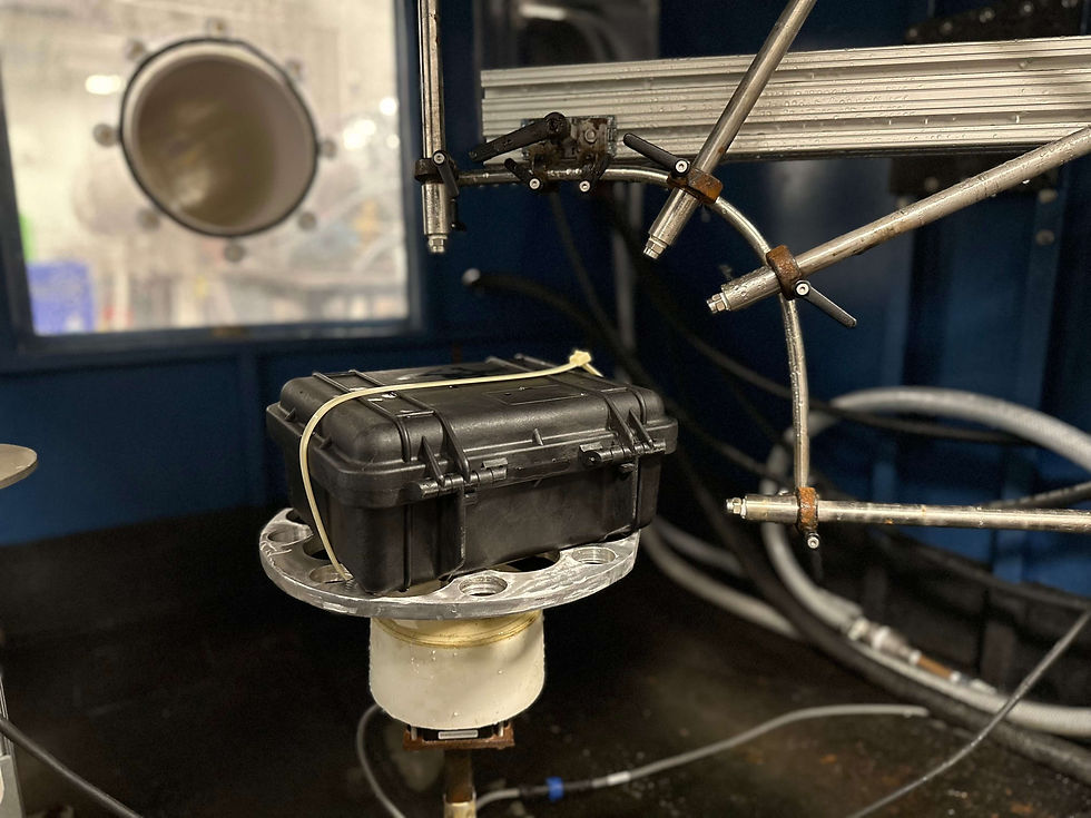 black plastic enclosure strapped to turntable inside a water ingress testing chamber, with IPX9k nozzles shown