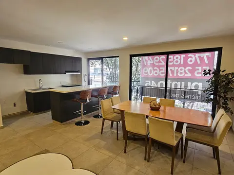 Área de comedor y cocina de concepto abierto, con mobiliario de madera, barra desayunadora oscura y ventanales de piso a techo con vista al exterior.