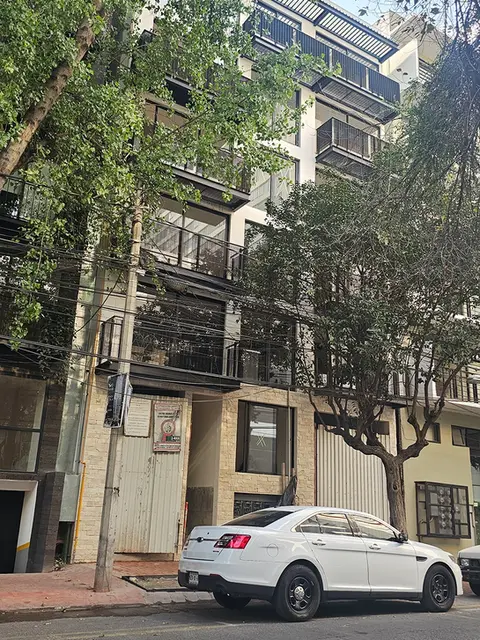 Fachada exterior de un edificio residencial moderno con balcones de herrería negra, amplios ventanales y vegetación urbana integrada en la entrada principal.