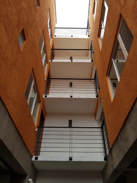 Perspectiva de pasillos internos en edificio residencial con muros naranja y techos blancos, destacando la iluminación natural y el diseño de herrería.