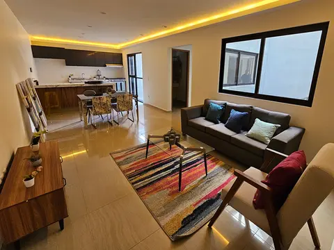 Sala de estar que destaca por su diseño de iluminación cálida indirecta en el plafón, creando un ambiente acogedor y sofisticado hacia el área de cocina.