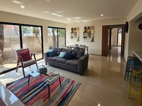 Estancia iluminada por grandes ventanales de piso a techo con vista a áreas verdes, decorada con una alfombra multicolor y mobiliario contemporáneo.