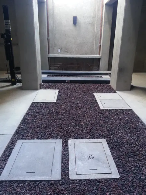 Detalle técnico de área de estacionamiento con piso de grava roja (tezontle), registros de concreto y base de sistema elevautos para ahorro de espacio.