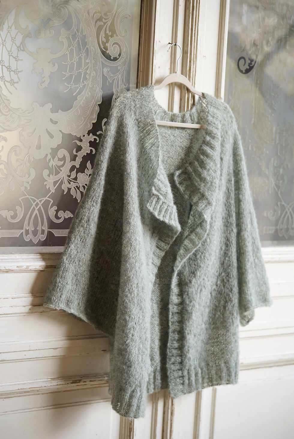 Szałwiowy kardigan ręcznie robiony na drutach widok przodu, wisi na wieszaku na tle drewnianych kremowych drzwi