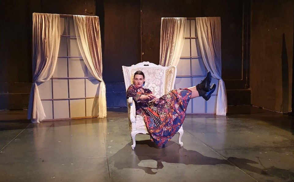 Η αφηγήτρια Κωνσταντίνα Δαούτη, καθισμένη με τα πόδια πάνω στην πολυθρόνα, στο κέντρο της σκηνής