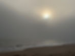 The sun glows dimly through thick coastal fog over a quiet beach, casting a soft golden light.