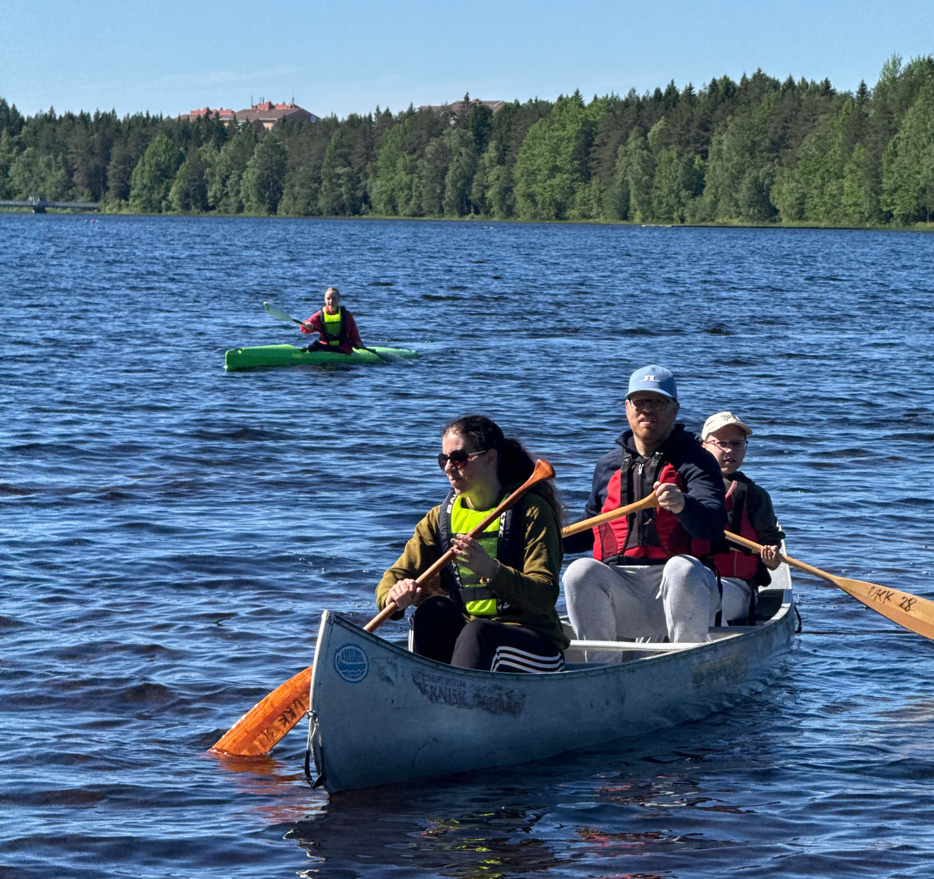 Wide-angle view of children and adults paddling kayaks near a calm Swedish lake shore.