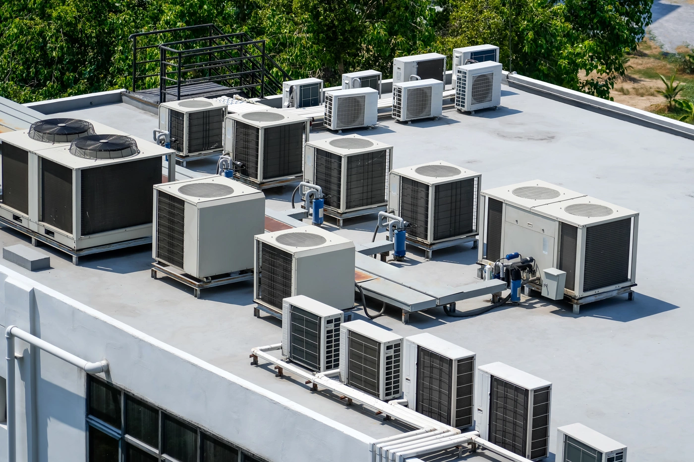 Numerous air conditioning units on a flat rooftop against a backdrop of green trees. White and gray colors dominate the scene.