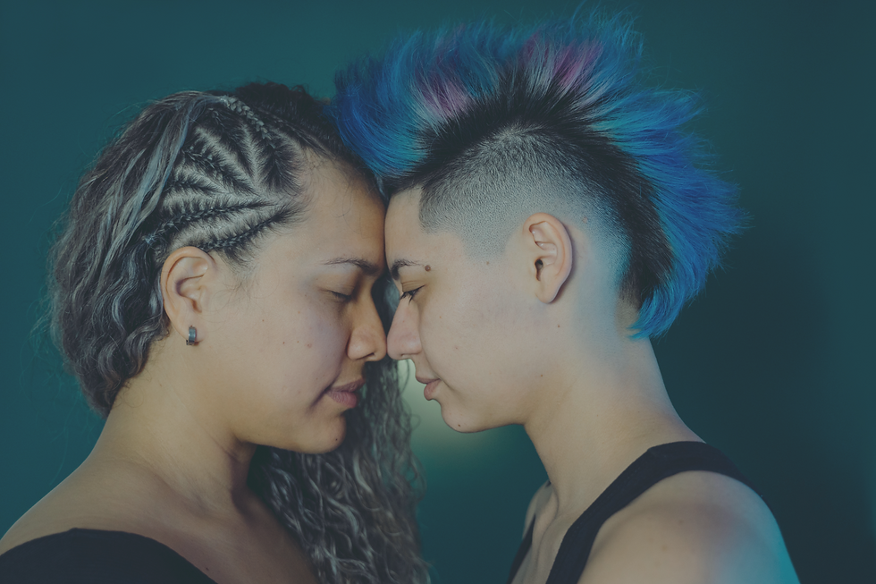 Two people touching foreheads with eyes closed, in a serene moment. Both have unique hairstyles; one sports blue and purple spikes.