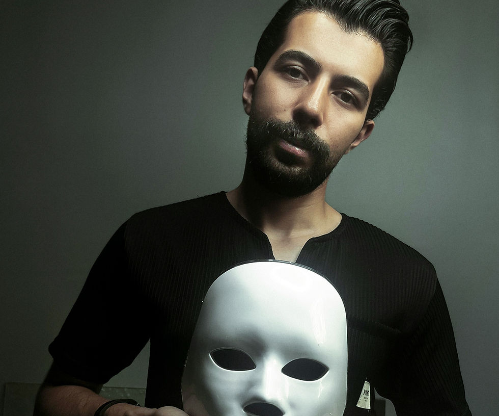 Man holding a white mask, wearing a black shirt in a dimly lit, neutral background. Mood is mysterious and contemplative.