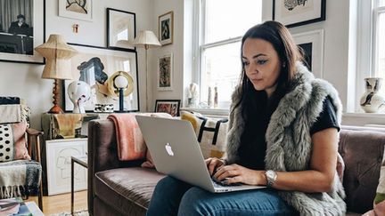 Lucy Machado, Woman, on a couch using a laptop in a cozy, art-filled living room with bright natural light. Various framed artworks and decor pieces adorn the walls.