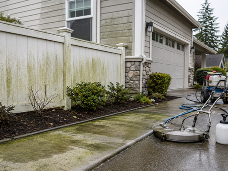 Algae-stained concrete walkway, fence, and siding with professional soft washing equipment staged outside a home.