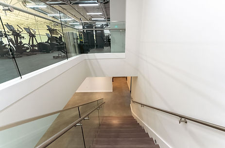 Interior staircase with glass railing connecting upper fitness level to lower floor in commercial sports facility.