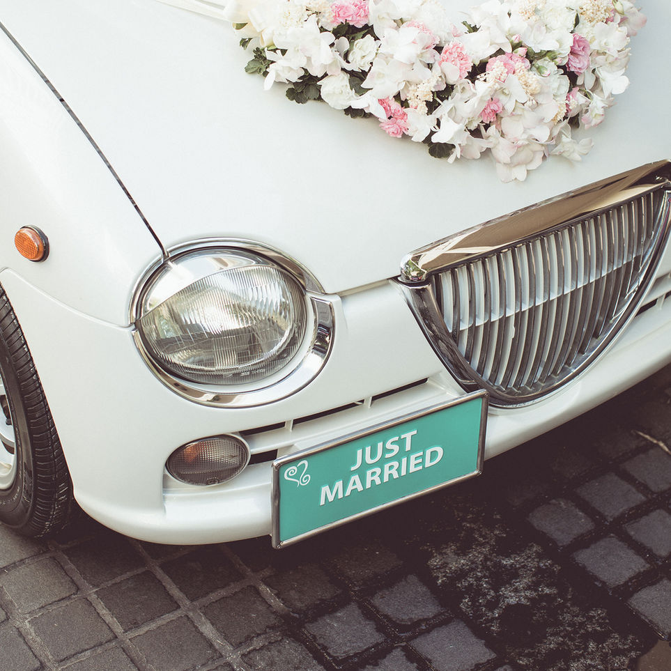 White vintage car with flower bouquet, "Just Married" sign on front. Car on cobblestone street, evoking joy and celebration.