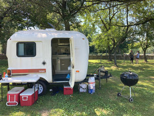 Camper, charcoal grill, coolers, and chairs at a campsite.