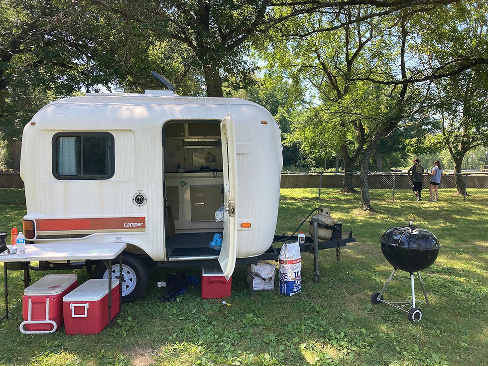 Camper, charcoal grill, coolers, and chairs at a campsite.