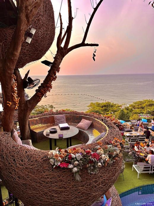 Twilight seaside rooftop lounge in a woven tree pod, with diners below and a calm ocean sunset in the background.