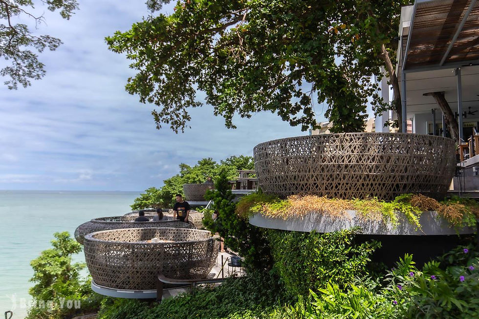 Seaside resort balconies with wicker pod seating overlook the ocean, a few people relaxing amid lush greenery; faint watermark bottom left.