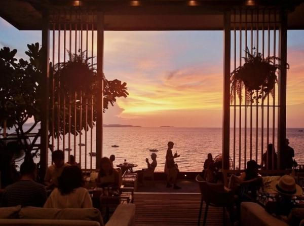 Silhouetted diners relax at a seaside restaurant patio, framed by hanging plants and a glowing orange-pink sunset over the ocean.