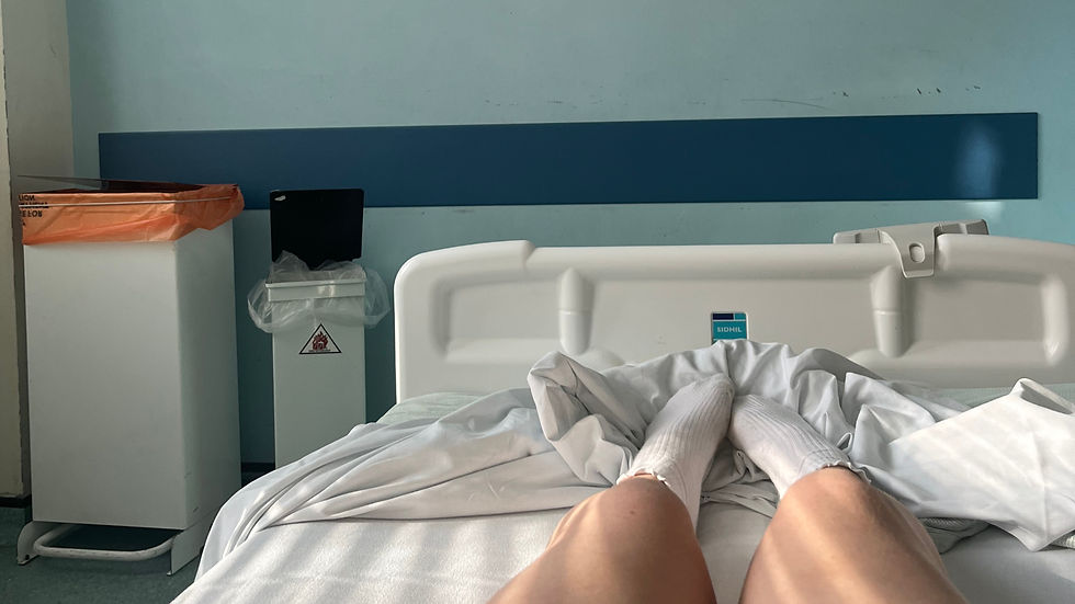 Person's legs in white socks on a hospital bed, room with blue wall. Trash bins on the left. Calm mood. Brand label "SIDHIL" visible.