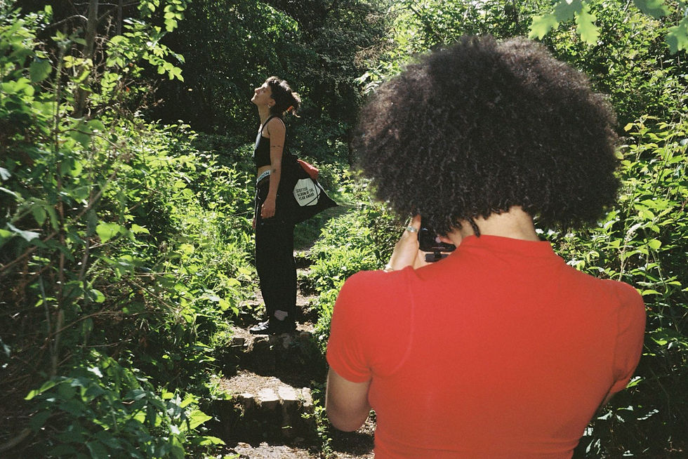 Two people in a lush, green forest; one in red photographing the other, who is looking up, relaxed. Sunlight filters through leaves.