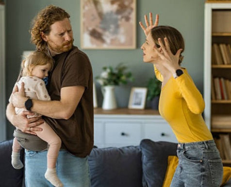 A couple argues in a living room; the man holds a baby. The woman gestures emphatically. The room is decorated in soft tones.